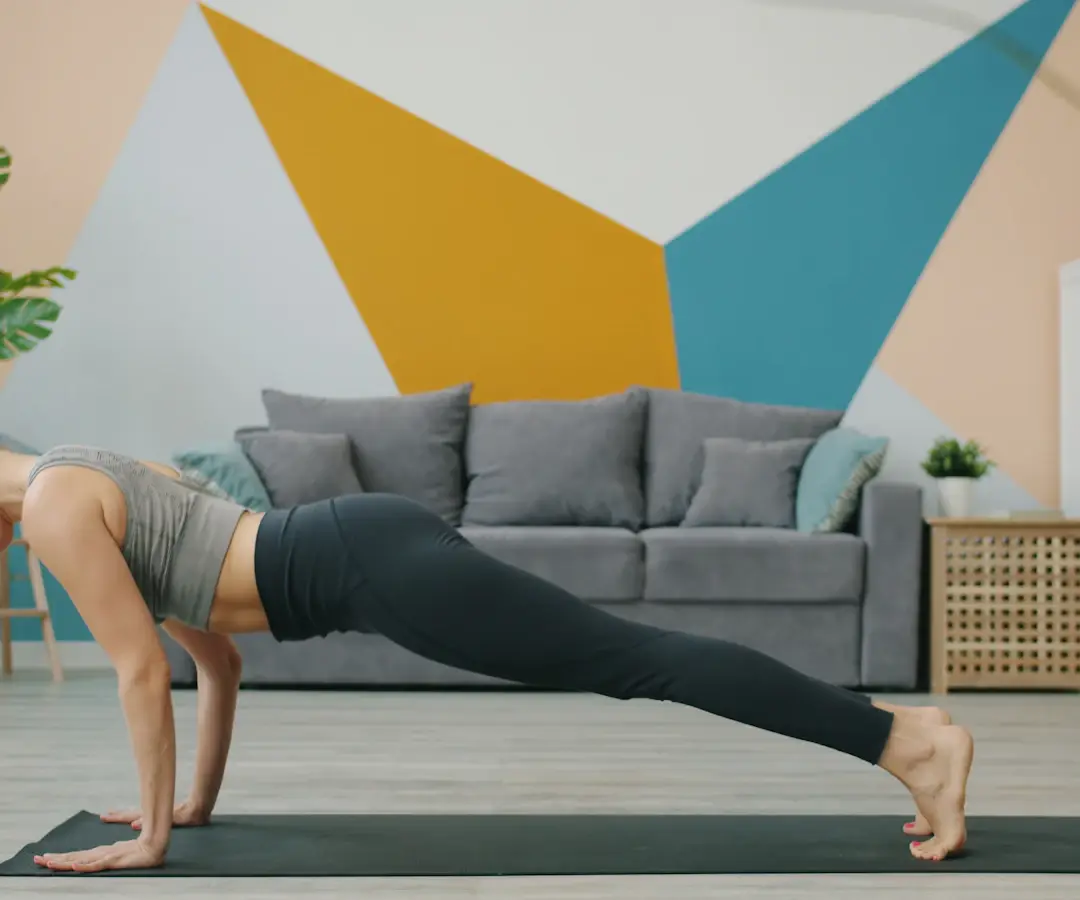 Femme en position de planche bras tendus sur un tapis, dans un salon au mur peint de triangles colorés