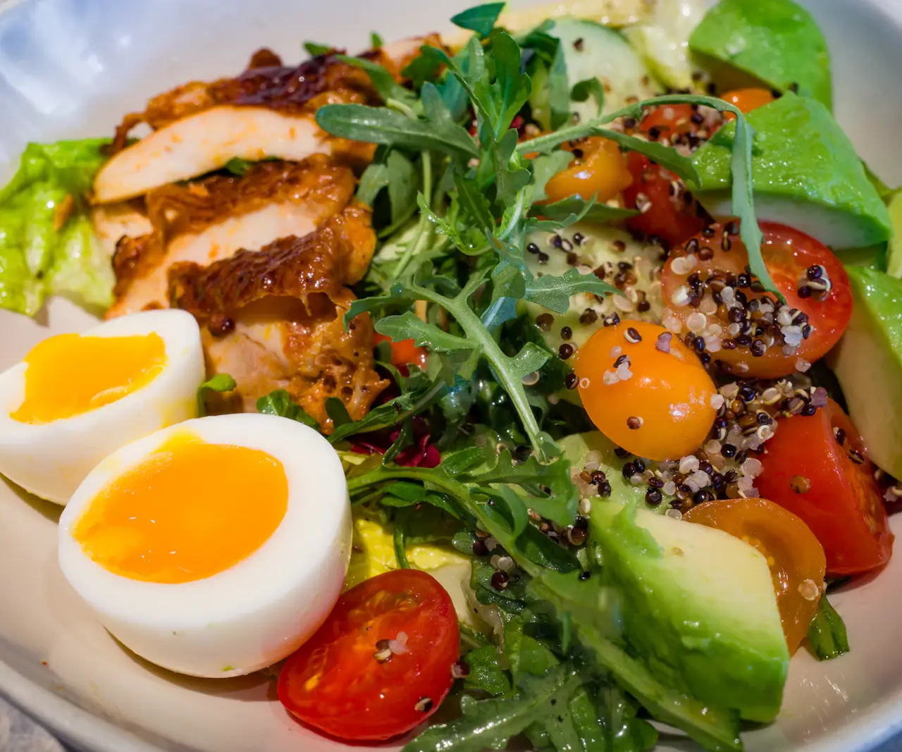 Bol de salade avec poulet grillé tranché, deux demi-oeufs mollets, avocat, tomates cerises, roquette et quinoa