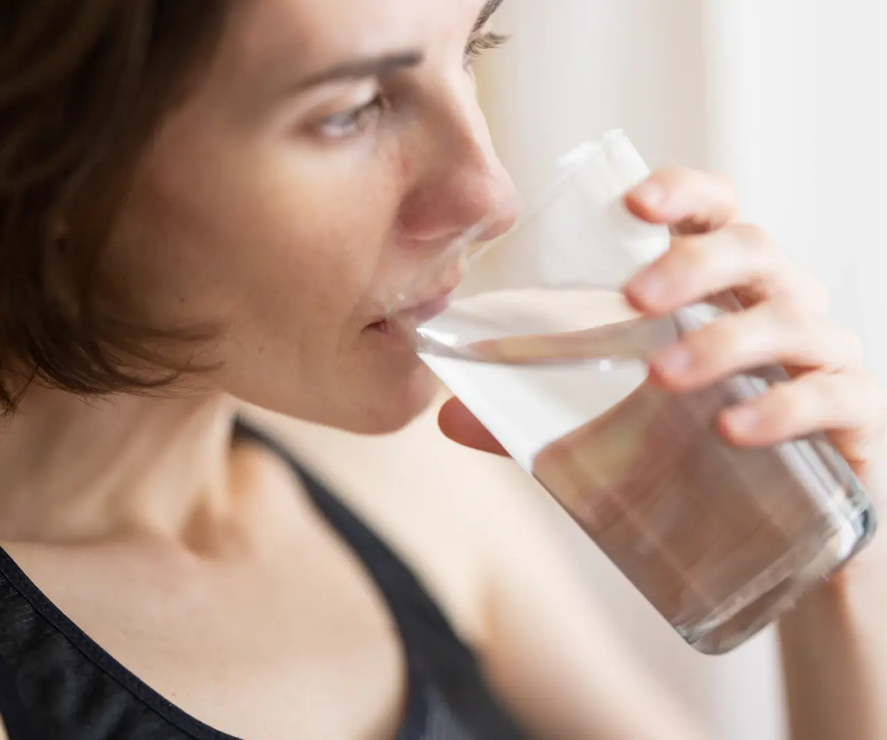 Femme aux cheveux courts en débardeur de sport noir buvant un grand verre d'eau près d'une fenêtre