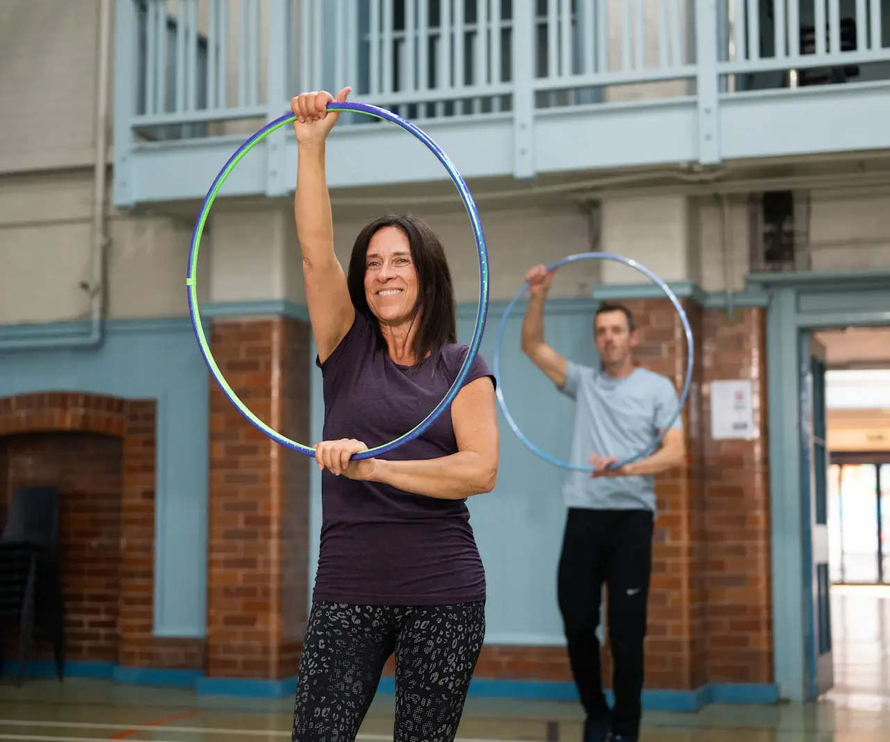 Femme souriante en t-shirt violet qui tient un cerceau au-dessus de sa tête dans une salle de sport, un homme fait de même derrière elle