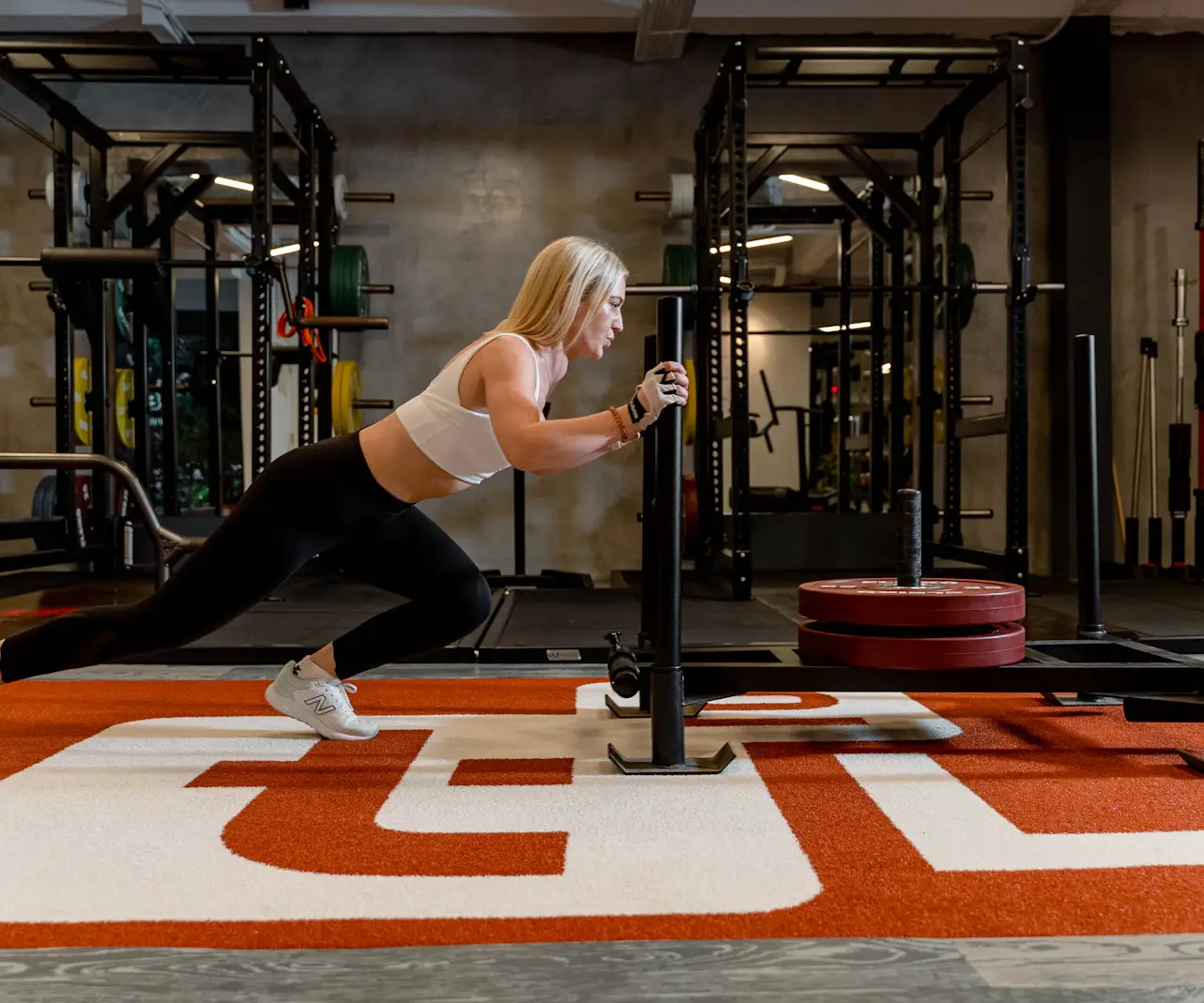 Femme blonde en brassière blanche qui pousse un traîneau chargé de disques rouges sur une piste de gazon synthétique, dans une salle de sport