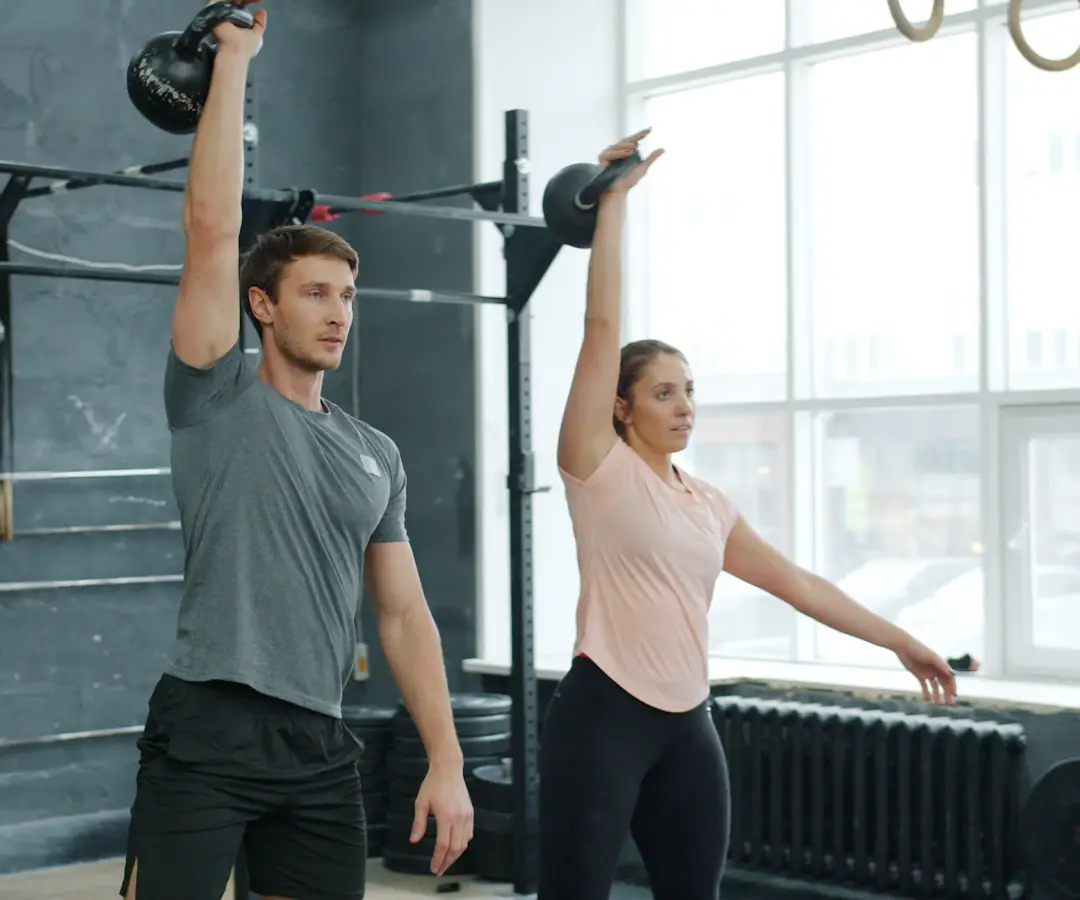 Un homme et une femme tiennent chacun une kettlebell bras tendu au-dessus de la tête dans une salle de sport lumineuse