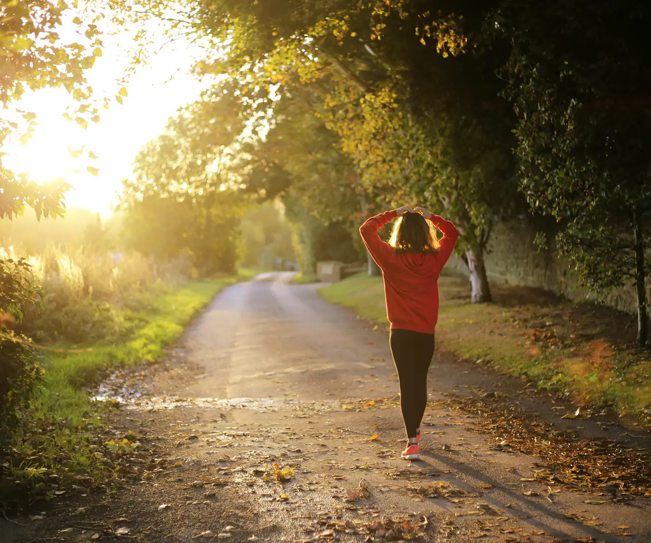 Femme de dos en sweat rouge qui marche sur un chemin bordé d'arbres, les mains sur la tête, dans la lumière dorée du soleil bas