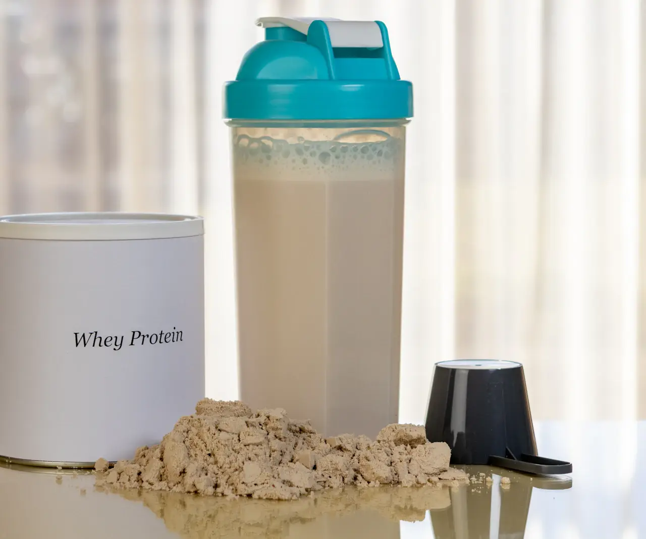 Pot de whey protéine blanc, shaker rempli d'une boisson protéinée, tas de poudre beige et doseur noir sur une table