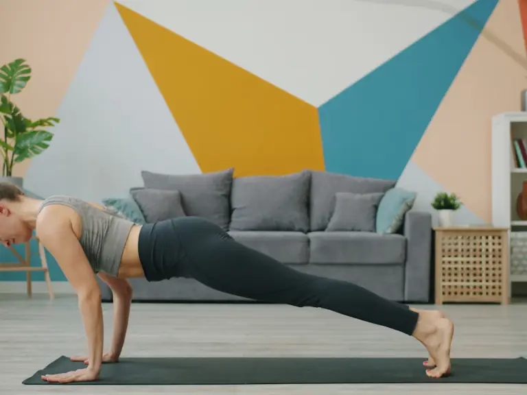 Femme en position de planche bras tendus sur un tapis, dans un salon au mur peint de triangles colorés