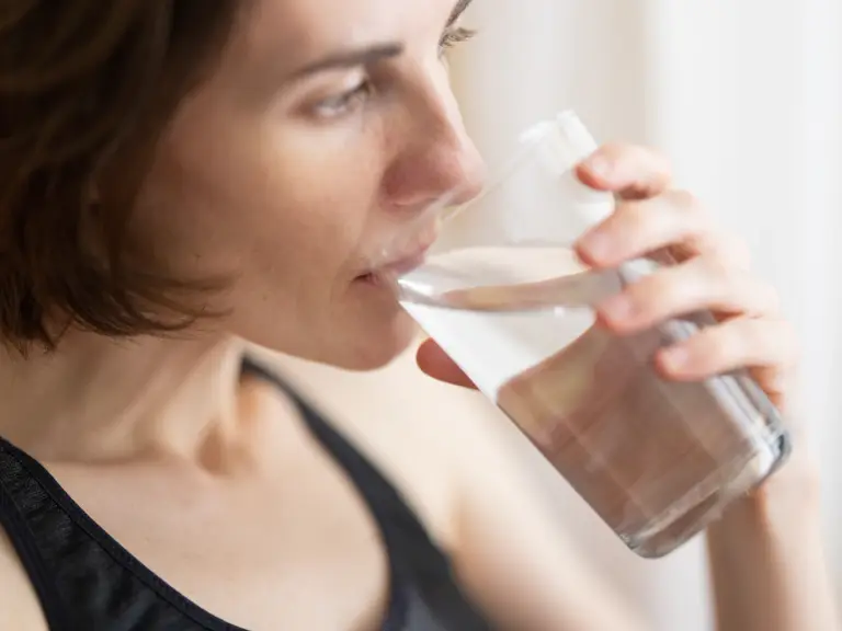 Femme aux cheveux courts en débardeur de sport noir buvant un grand verre d'eau près d'une fenêtre