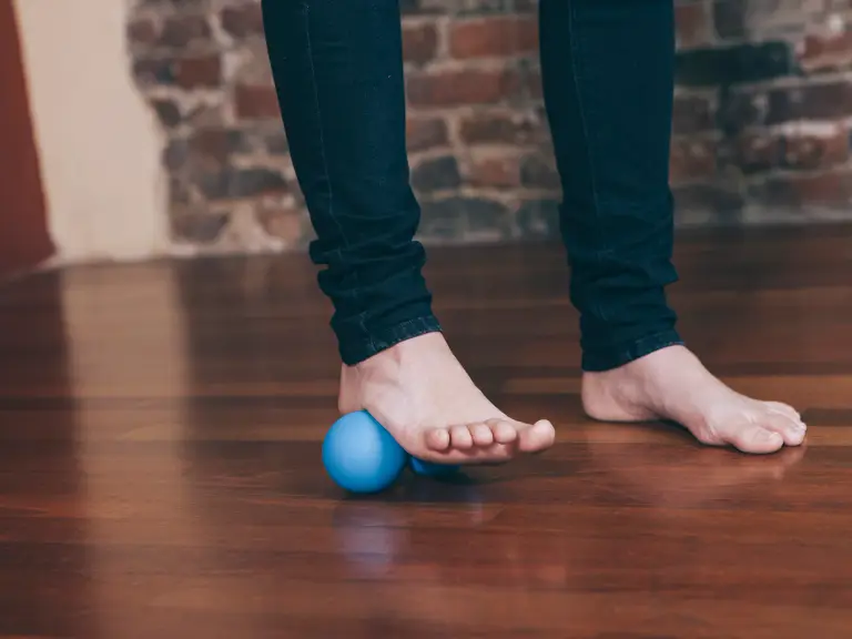 Pieds nus sur un parquet, un pied qui roule sur une balle de massage bleue pour détendre la voûte plantaire