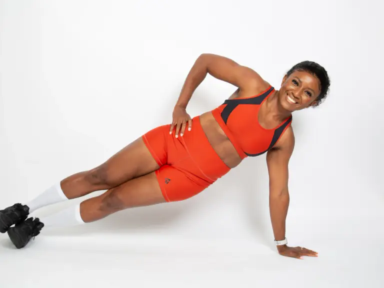 Femme souriante en tenue de sport orange qui tient une planche latérale sur une main, sur fond blanc