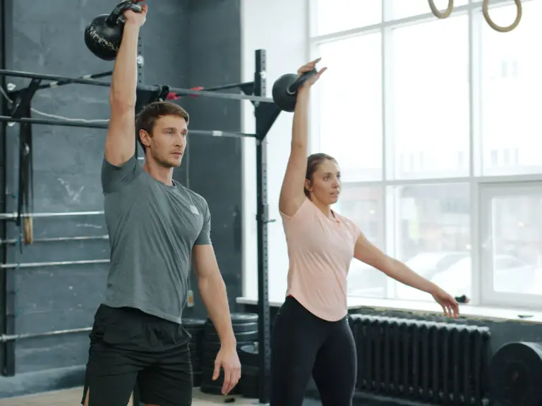 Un homme et une femme tiennent chacun une kettlebell bras tendu au-dessus de la tête dans une salle de sport lumineuse