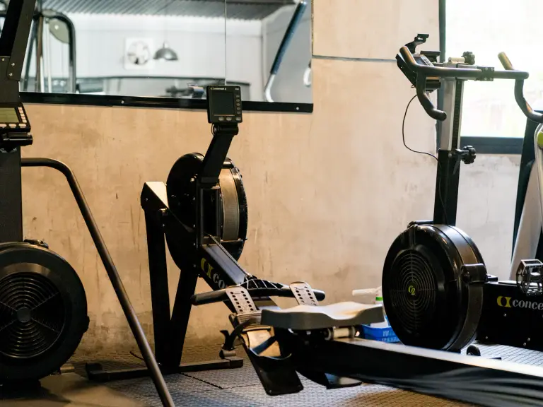 Rameur à air posé entre deux machines de cardio, dont un vélo à volant d'inertie, contre un mur en béton