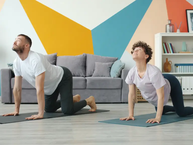 Un homme et une femme à quatre pattes sur des tapis de yoga dans un salon, en train de faire l'exercice du chat-vache