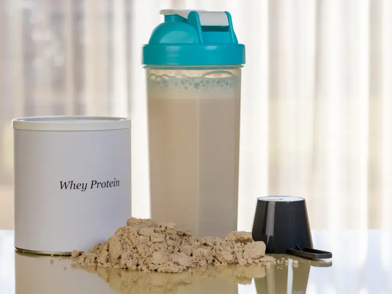 Pot de whey protéine blanc, shaker rempli d'une boisson protéinée, tas de poudre beige et doseur noir sur une table