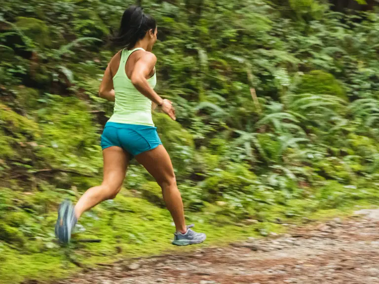 Femme en débardeur vert clair et short bleu qui court sur un chemin de terre en forêt